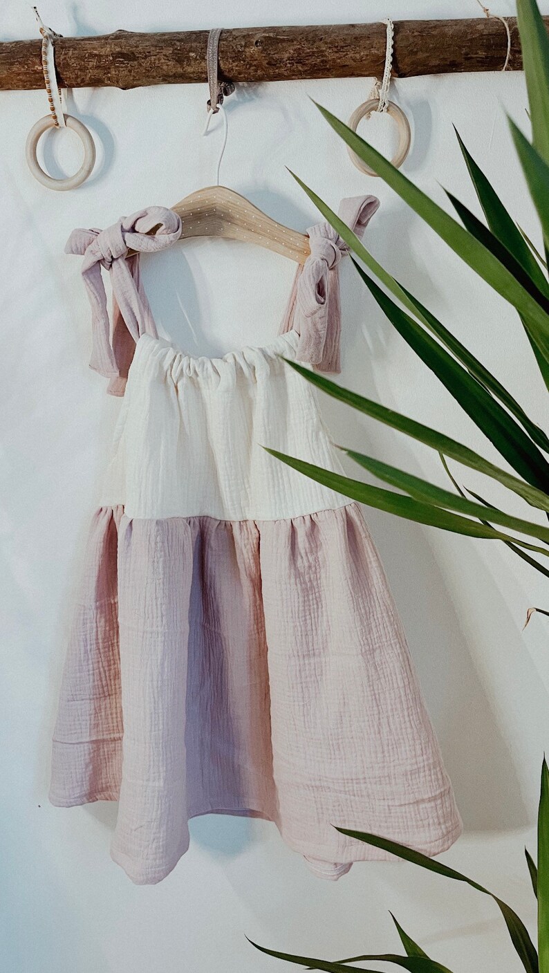 May include: A white and pink sleeveless dress with a gathered bodice and a ruffled hem. The dress is hanging on a wooden hanger with a pink ribbon tied around the straps.