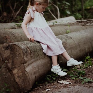 May include: A young girl wearing a pink and white dress sits on a large log in a forest setting. She is wearing white sneakers and has her hair pulled back in a ponytail.