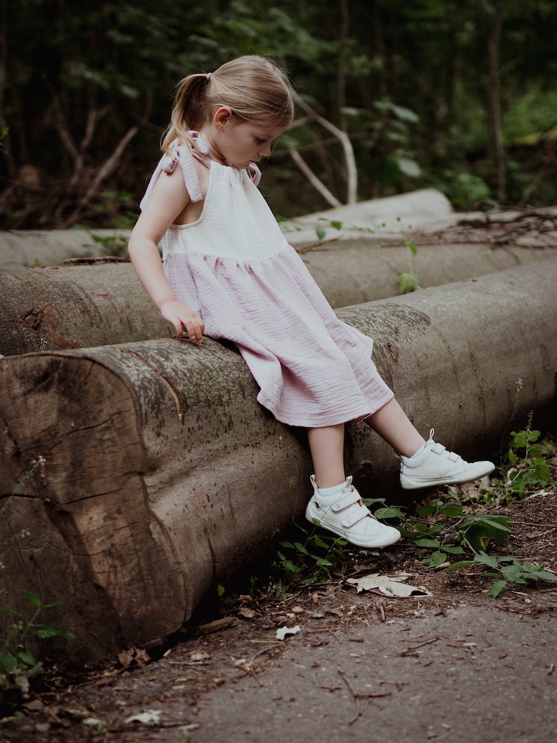 May include: A young girl wearing a pink and white dress sits on a large log in a forest setting. She is wearing white sneakers and has her hair pulled back in a ponytail.