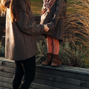 May include: Two people, one wearing a brown coat and the other wearing a brown sweater and pink leggings, stand on a wooden platform. The person in the coat is looking at the person in the sweater, who is smiling.