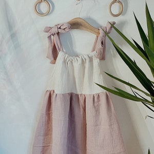 May include: A pink and white sleeveless dress with a gathered skirt and knotted straps. The dress is hanging on a wooden hanger with two wooden rings on a branch above.