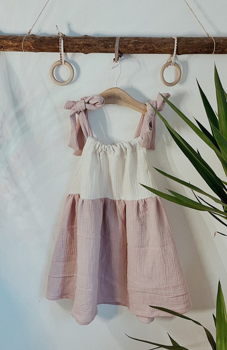 May include: A pink and white sleeveless dress with a gathered skirt and knotted straps. The dress is hanging on a wooden hanger with two wooden rings on a branch above.
