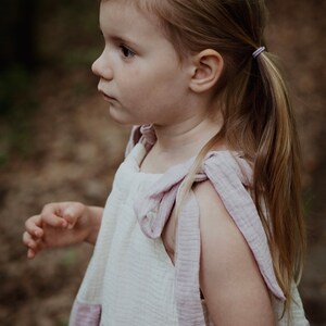 May include: A young girl with long blonde hair wearing a white and pink dress with a bow on the strap. She is standing in a forest setting.