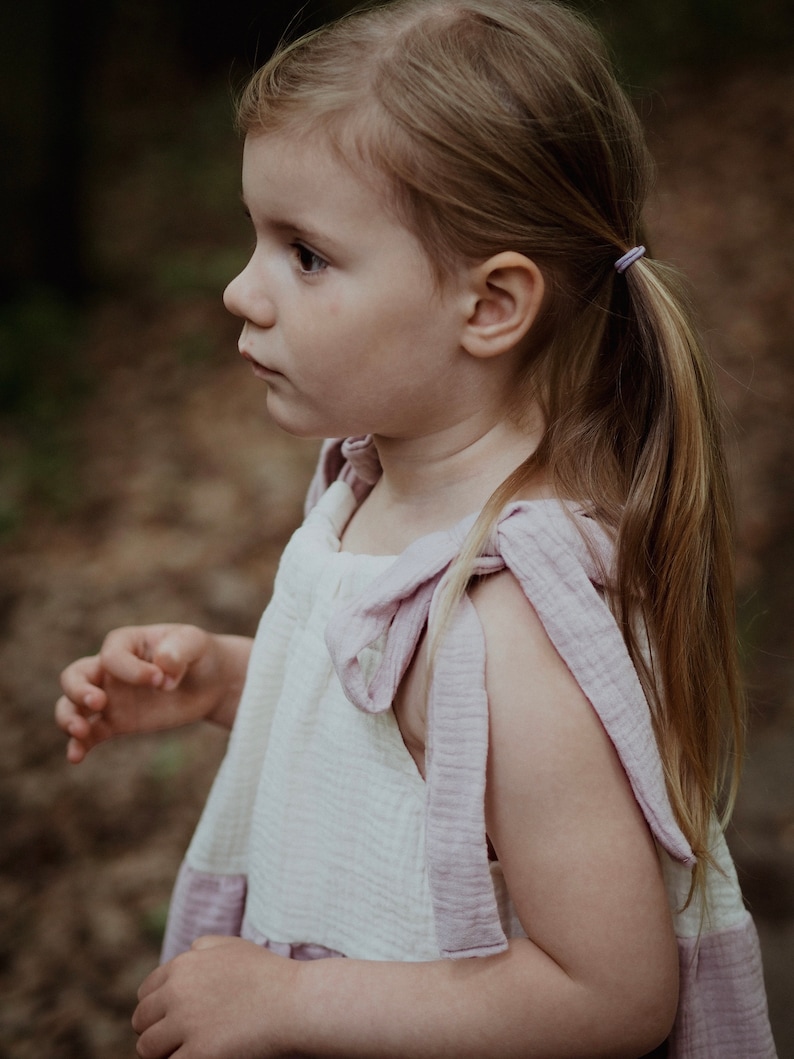 May include: A young girl with long blonde hair wearing a white and pink dress with a bow on the strap. She is standing in a forest setting.