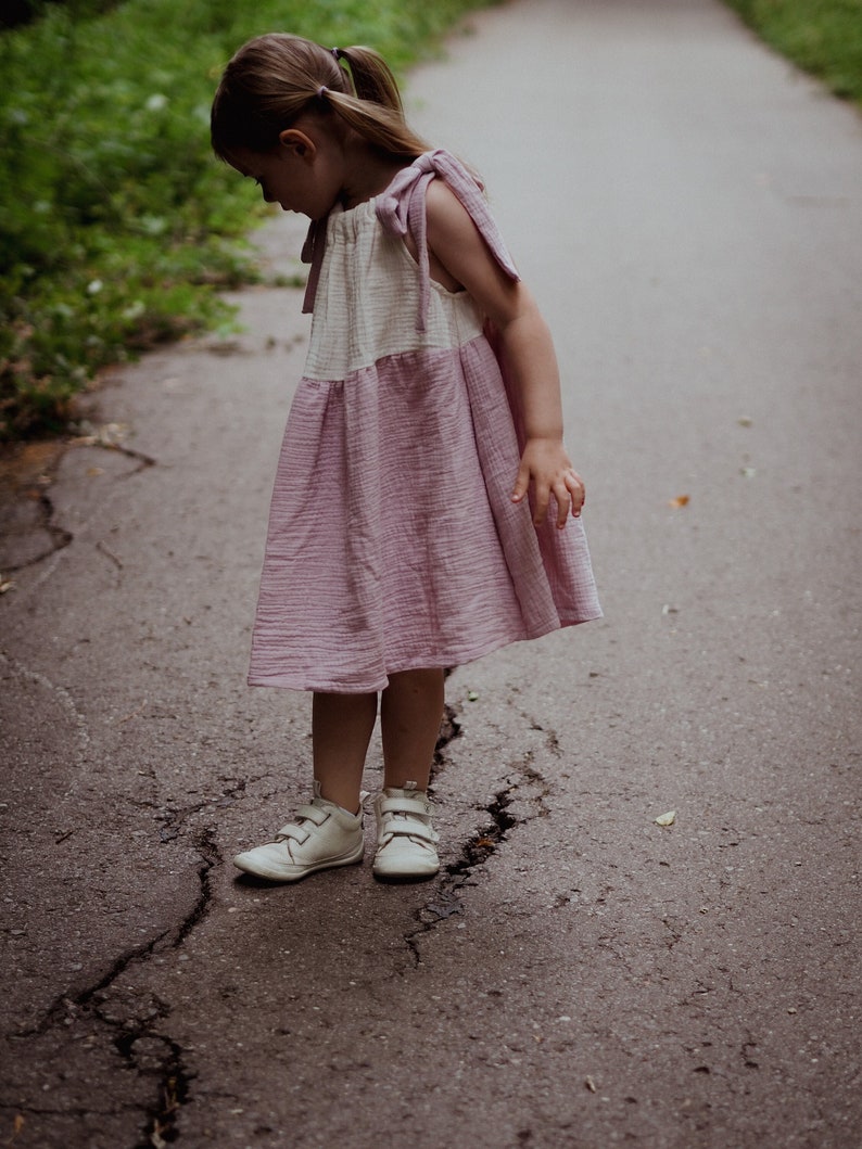 May include: A young girl wearing a pink and white dress with a ruffled hem and white sneakers stands on a cracked asphalt path.
