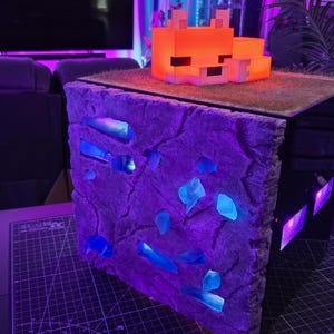 Può includere: Una lampada a tema Minecraft con un design a fiamma arancione pixelato sopra una struttura a blocchi. Il blocco ha un esterno strutturato, viola-grigio con accenti di cristallo blu brillante. La lampada è su un tavolo.
