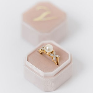 Pode incluir: Um anel de ouro com uma pérola e pequenos diamantes engastados em um delicado design floral. O anel é exibido em uma caixa de anel de veludo rosa claro.