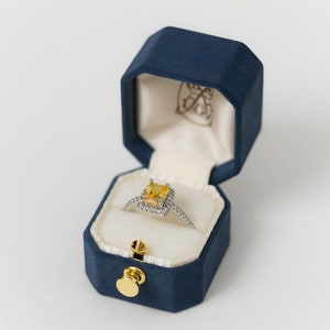 May include: A yellow gemstone ring with a halo setting, set in a blue velvet ring box.