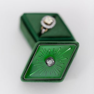 Puede incluir: Un joyero de terciopelo verde con tapa en forma de diamante con una gema gris central y líneas radiales. La caja está abierta, revelando un anillo con una piedra central grande y clara y un halo de piedras más pequeñas.