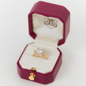 Puede incluir: Un anillo de compromiso de oro con una piedra central grande y transparente y piedras más pequeñas alrededor, en un diseño floral. El anillo se muestra en un joyero octogonal burdeos abierto, con interior blanco y cierre dorado.