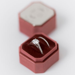 Puede incluir: Un anillo de compromiso de diamantes en un joyero de terciopelo rosa. El anillo presenta un diamante grande de talla redonda y una banda delicada con piedras de acento más pequeñas. Se ve un segundo anillo.