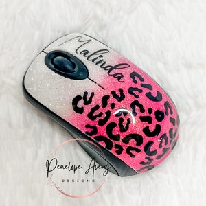 May include: A wireless computer mouse featuring a glittery white to pink gradient finish with a black leopard print pattern. The name Malinda is printed in black script on the top surface, and the logo Penelope Avery DESIGNS is visible.