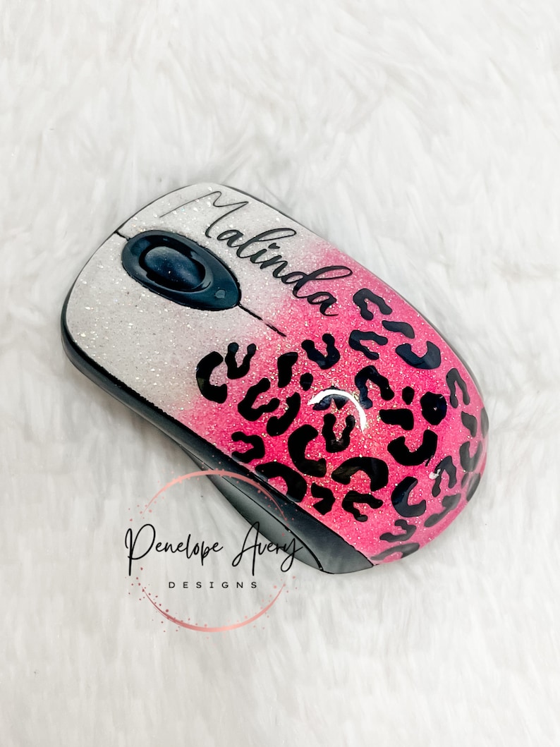 May include: A wireless computer mouse featuring a glittery white to pink gradient finish with a black leopard print pattern. The name Malinda is printed in black script on the top surface, and the logo Penelope Avery DESIGNS is visible.
