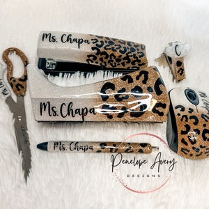 May include: A set of office supplies including scissors, a stapler, a tape dispenser, a pen, a computer mouse, and a small clip, all featuring a gold glitter and black leopard print pattern with the name Ms. Chapa printed on each item.