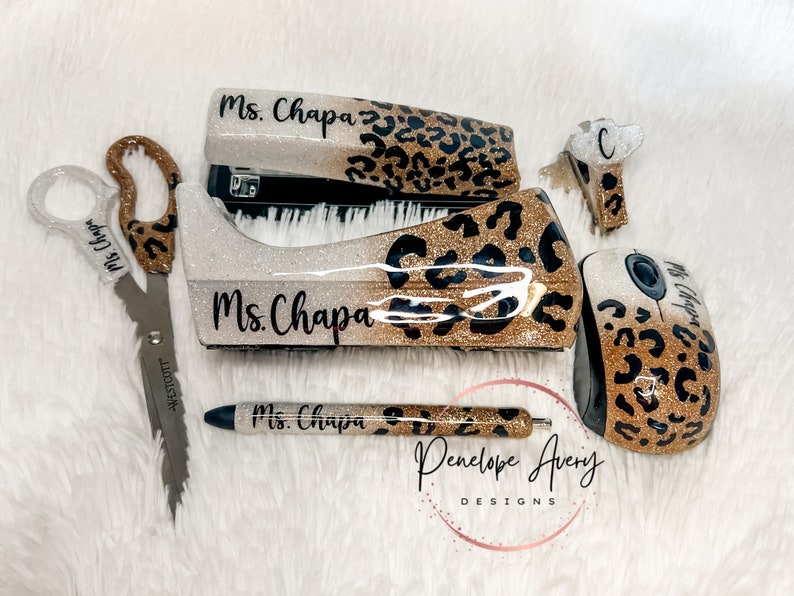 May include: A set of office supplies including scissors, a stapler, a tape dispenser, a pen, a computer mouse, and a small clip, all featuring a gold glitter and black leopard print pattern with the name Ms. Chapa printed on each item.