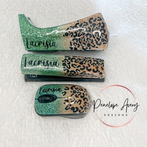 May include: A stapler, a tape dispenser, and a computer mouse, all featuring a glittery green and gold leopard print finish. Each item is personalized with the name Lacrisia in black script lettering. The items are arranged on a white surface.