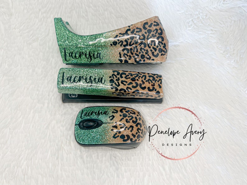 May include: A stapler, a tape dispenser, and a computer mouse, all featuring a glittery green and gold leopard print finish. Each item is personalized with the name Lacrisia in black script lettering. The items are arranged on a white surface.