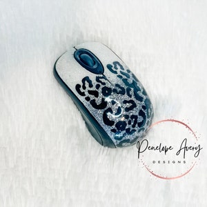 May include: A wireless computer mouse featuring a silver glitter finish with a black leopard print pattern. The device includes a central black scroll wheel and a black base. The text Penelope Avery DESIGNS is visible in the lower right corner.