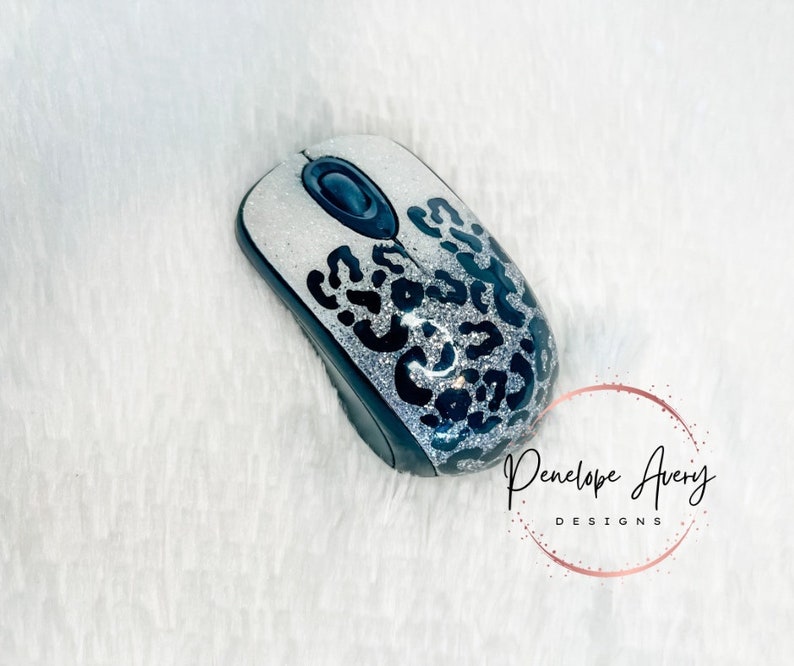 May include: A wireless computer mouse featuring a silver glitter finish with a black leopard print pattern. The device includes a central black scroll wheel and a black base. The text Penelope Avery DESIGNS is visible in the lower right corner.
