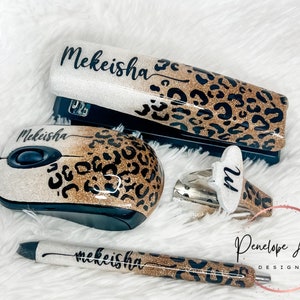 May include: A stapler, computer mouse, staple remover, and pen set featuring a glittery gold and white ombre finish with a black leopard print pattern. Each item is personalized with the name Mekeisha in black script, except the staple remover.