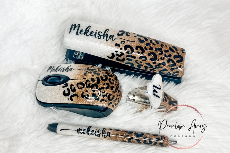 May include: A stapler, computer mouse, staple remover, and pen set featuring a glittery gold and white ombre finish with a black leopard print pattern. Each item is personalized with the name Mekeisha in black script, except the staple remover.