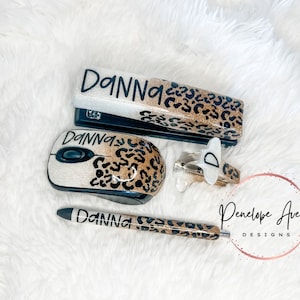 May include: A stapler, computer mouse, pen, and staple remover set featuring a white and gold glitter finish with a black leopard print pattern. Each item is personalized with the name "DANNA" in black script, except for the staple remover.