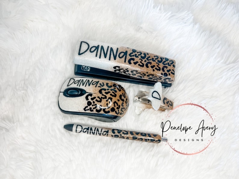 May include: A stapler, computer mouse, pen, and staple remover set featuring a white and gold glitter finish with a black leopard print pattern. Each item is personalized with the name "DANNA" in black script, except for the staple remover.