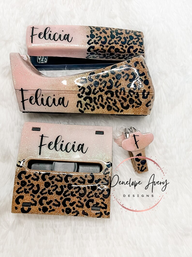 May include: A desk set featuring a stapler, tape dispenser, and note holder, all finished in a pink and gold glitter gradient with a black leopard print pattern. Each item is personalized with the name Felicia in black script lettering.