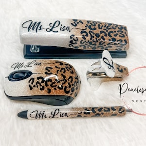 May include: A custom office set featuring a stapler, a computer mouse, a staple remover, and a pen. Each item has a white-to-gold glitter gradient finish with a black leopard print pattern and the name Ms. Lisa printed in black script lettering.