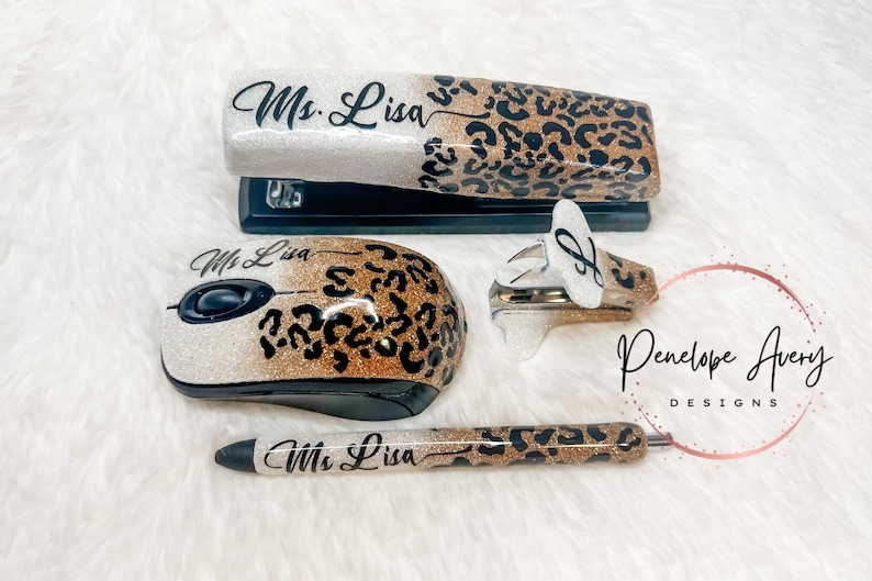 May include: A custom office set featuring a stapler, a computer mouse, a staple remover, and a pen. Each item has a white-to-gold glitter gradient finish with a black leopard print pattern and the name Ms. Lisa printed in black script lettering.
