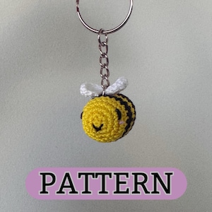 May include: A crocheted bee keyring with black and white stripes, yellow body, and white wings. The bee has a smiling face and is attached to a silver key ring.