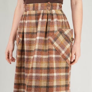 May include: A vintage plaid skirt in shades of brown, tan, and cream. The skirt features a high waist, pleats, and two front pockets. The fabric appears to be a woven material, and the skirt falls to the mid-calf.