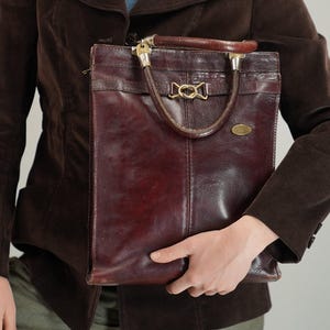 Könnte beinhalten: Eine Vintage-Tote-Tasche aus weinrotem Leder mit einem oberen Griff und goldfarbener Hardware. Die Tasche hat eine rechteckige Form und ein strukturiertes Design. Der Griff ist aus braunem Leder. Die Tasche wird vor einer Person gehalten, die eine braune Jacke und ein Jeanshemd trägt.