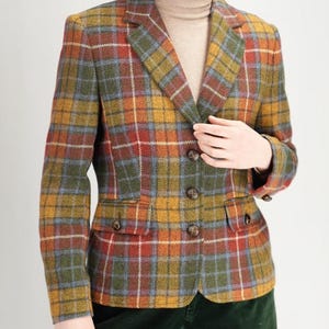 Könnte beinhalten: Ein mehrfarbiger Blazer mit Karomuster, Knopfverschluss und zwei Vordertaschen. Der Blazer weist eine Mischung aus roten, gelben, grünen und blauen Farbtönen auf. Die Person trägt einen beigen Rollkragenpullover und eine dunkelgrüne Hose.
