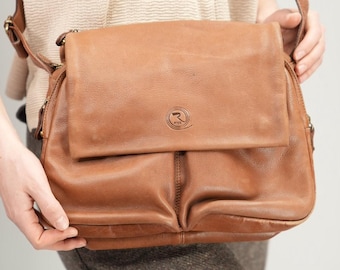 May include: A brown leather messenger bag with a flap closure and adjustable shoulder strap. The bag has a logo embossed on the front and is held by a person. The bag is made of a soft, worn leather.