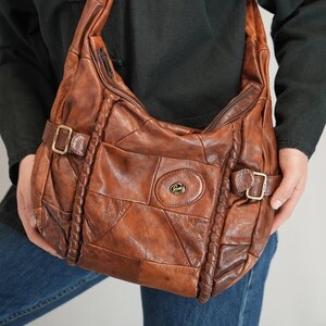 May include: A brown leather shoulder bag with a patchwork design. The bag features a long adjustable strap, a zippered top, and decorative braided accents. It has two buckle closures and a small logo on the front.