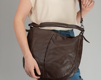 May include: A dark brown leather hobo bag with a smooth finish, featuring a single wide shoulder strap, a curved silhouette, and a central vertical seam detail. The bag is worn over the shoulder with a long adjustable strap attached by silver metal hardware.