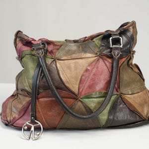 Könnte beinhalten: Eine große Patchwork-Lederhandtasche in Braun-, Grün- und Rottönen. Die Tasche hat zwei Griffe, einen silberfarbenen Peace-Zeichen-Anhänger und eine silberne Schnalle. Die Tasche steht auf einer weißen Oberfläche.