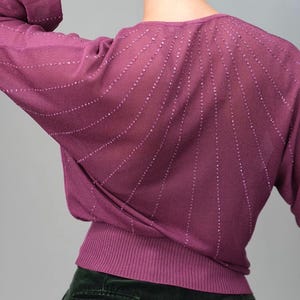 Può includere: Un maglione color malva con orlo a coste e maniche lunghe. La parte posteriore del maglione presenta un disegno di linee verticali con piccoli abbellimenti scintillanti. La modella indossa pantaloni verde scuro.