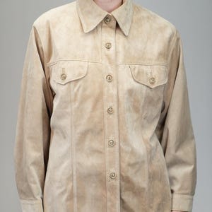 May include: A tan suede shirt with long sleeves, two chest pockets, and button fastenings. The shirt has a collared neckline and is buttoned up. The garment is worn with black trousers.