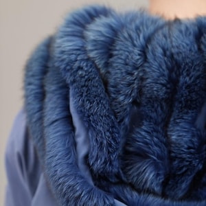 Op de afbeelding: Een blauwe capuchonjas met een bontkraag rond de capuchon. De bont is diepblauw van kleur en is erg zacht en pluche.