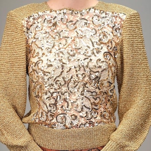 Può includere: Un maglione dorato con paillettes, maniche lunghe e scollo rotondo. Il maglione presenta un delicato motivo a pizzo con paillettes dorate su tutta la superficie. Il maglione è abbinato a una gonna paisley marrone e nera.