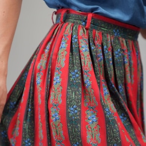 May include: A red and green striped skirt with a floral pattern. The skirt has vertical stripes of red and green, with a repeating design of blue flowers and green leaves. The skirt is made of a lightweight fabric.