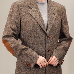 Puede incluir: Una chaqueta de tweed marrón con parches de cuero en los codos y botones marrones. La chaqueta tiene un cierre de un solo botón y una solapa de muesca.