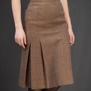 Pode incluir: Saia de tweed castanho com frente plissada. A saia tem comprimento até ao joelho e apresenta um padrão xadrez subtil. A modelo usa collants pretas e um top verde-azulado.