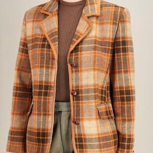 May include: A plaid blazer in shades of brown, orange, and tan. The jacket has a classic design with a notched collar, two front pockets, and brown buttons. The edges are trimmed with a lighter brown material. The person is wearing a brown sweater and green pants.