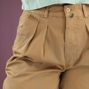 Könnte beinhalten: Eine braune, gefaltete Hose mit einem grünen Knopf und einer grünen Schlaufe an der Vorderseite. Die Hose ist aus einem weichen, bequemen Stoff.