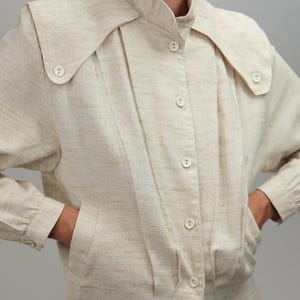May include: Cream-coloured button-up jacket with an unusual collar. The jacket has a high collar that extends over the shoulders, with buttons on the collar and down the front. It has long sleeves and pockets. The fabric has a subtle textured appearance.