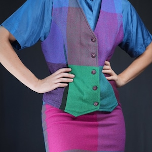 Op de afbeelding: Een geruit vest met een blauwe, paarse, groene en roze kleurstelling. Het vest heeft een knoopsluiting en wordt gedragen over een lichtblauw shirt. De persoon draagt een bijpassende geruite rok met een roze, blauwe en groene kleurstelling.