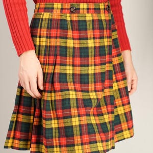 May include: A vintage tartan plaid skirt in red, yellow, and green. The skirt has a pleated design and is paired with a red long-sleeve top. The skirt is secured with two buttons at the waist. The skirt's pattern is a classic tartan design.